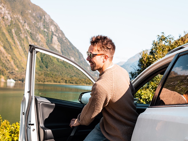 Smiling man stepping out of a car by a scenic lake with mountains in the background