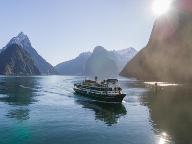 Cruise ship sailing through Milford Sound with snow-capped mountains and sunlight in the background